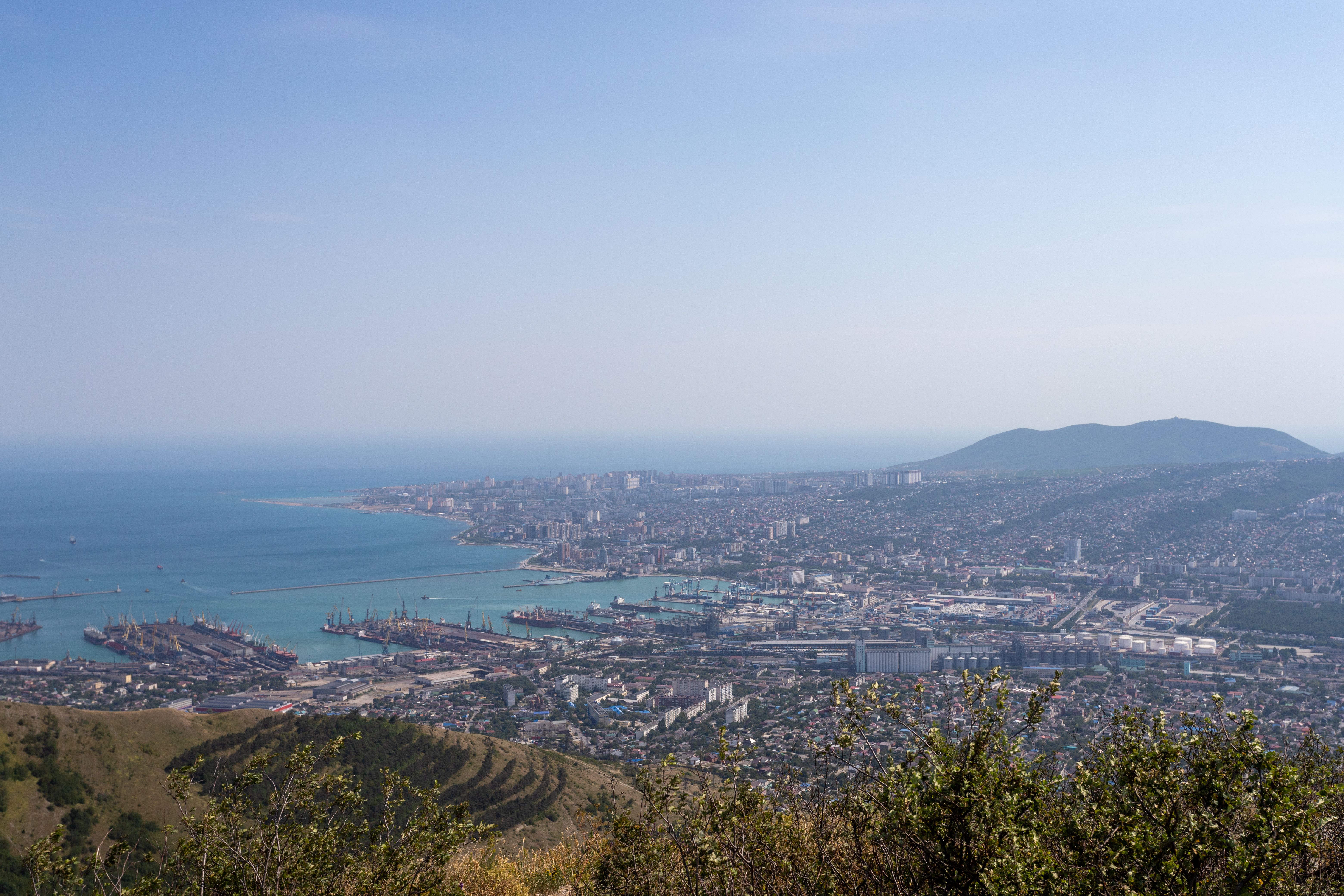 קרסנודאר: A scenic aerial view of Novorossiysk, Russia, highlighting the harbor against a backdrop of sea and hills.