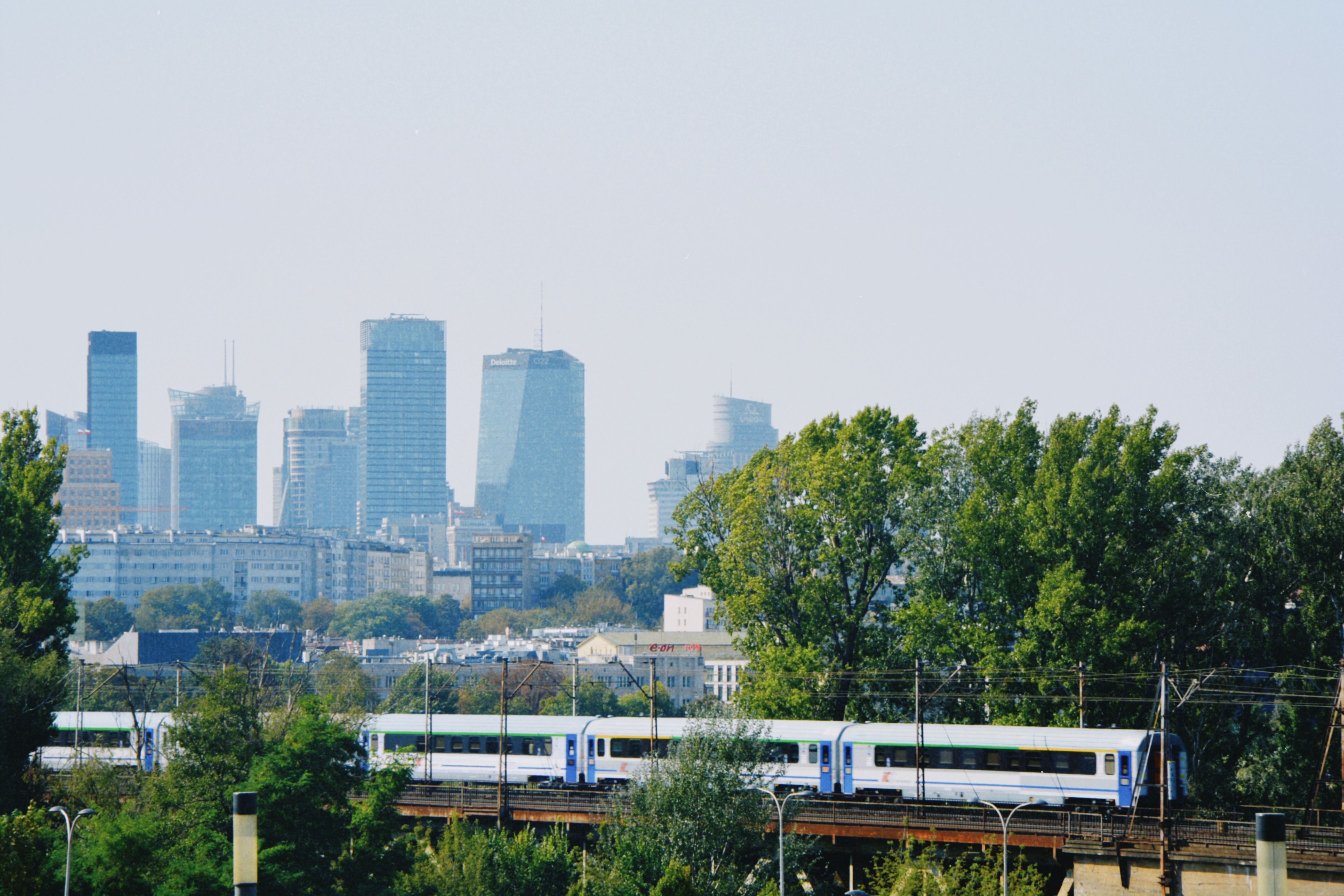 ורשה: Skyline of Warsaw with modern buildings and a train passing amidst the greenery.