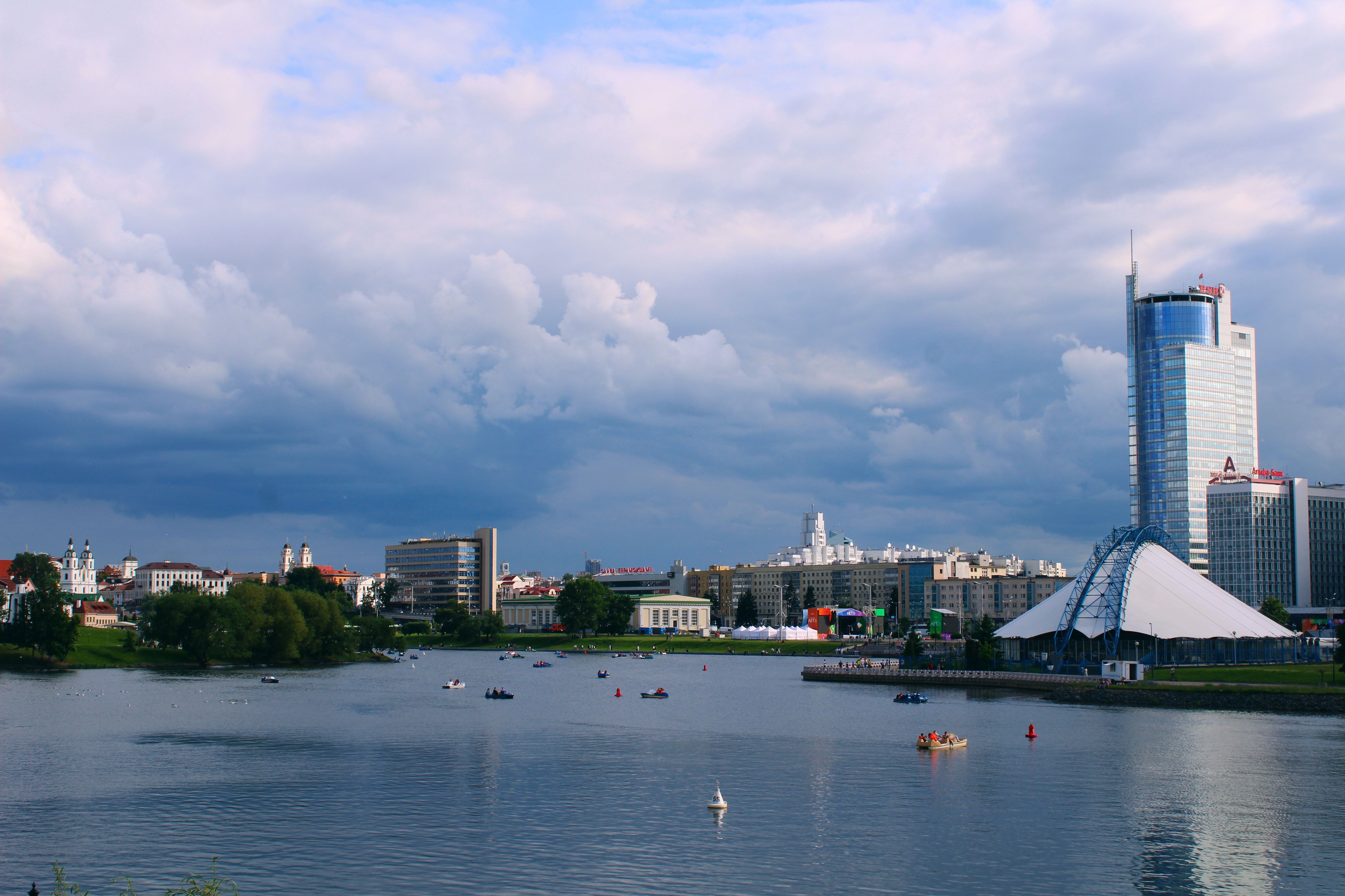מינסק: Picturesque view of Minsk cityscape along the river under a cloudy sky with modern and historic architecture.
